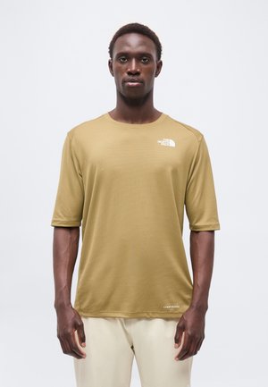 Homme debout face à l’avant, portant un t-shirt beige à manches courtes The North Face et un pantalon clair sur fond blanc.