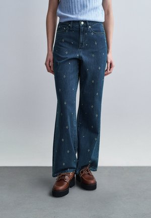 Kvinde iført højttaljede mørkeblå jeans med små lyse mønstre, kombineret med brune snørebåndssko med chunky såler.