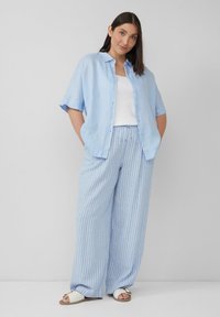 Lichtblauw overhemd met knopen over een witte tanktop, gecombineerd met brede blauw en witte gestreepte broek. Witte sandalen maken de outfit compleet.