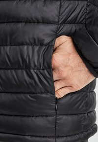 Chaqueta acolchada negra con un bolsillo con cremallera. El material es suave con un patrón acolchado y un ajuste ceñido alrededor del cuerpo.