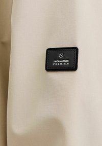 Čierna obdĺžniková nášivka Jack & Jones Premium prišitá na béžovej látke, zobrazujúca logo značky a text.