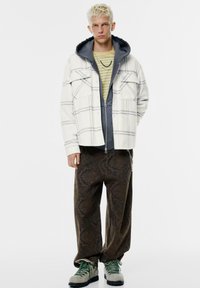 Kockovaný flanelový kabát v krémovej farbe s šedým vzorom, vrstvený cez šedú mikinu a pruhované zelené tričko, v kombinácii s hnedými širokými nohavicami so vzorom a šedými čižmami.