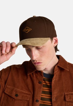 Jonge man die een bruine en beige pet verstelt, draagt een bruine corduroy jas over een gestreept shirt, kijkt met een neutrale uitdrukking naar beneden.