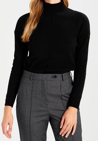 Schwarzer Strick-Turtleneck-Pullover mit strukturiertem Rippmuster, versehen mit einer Naht vorne, kombiniert mit grauen karierten Hochbundhosen.