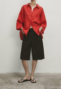 Chemise rouge en lin avec boutons, coupe décontractée, associée à un short marron foncé coupé au genou et des tongs noires. Design simple et décontracté.