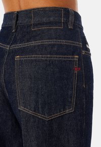 Detail kapsy na zadní straně tmavě modrých džín s oranžovým prošitím a malou červenou štítkovou značkou na pravé straně kapsy.