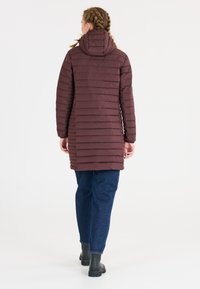Maroon puffy jakke med hætte, der har horisontale quiltede linjer. Båret over blå jeans og sorte støvler, set bagfra.