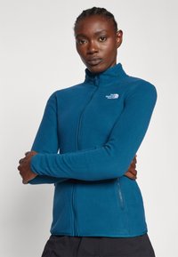 Chaqueta de forro polar en azul verdoso, con un cuello alto, cierre de cremallera y un pequeño logo en el pecho. Textura suave y diseño ajustado.