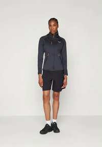 Naisen ulkoilutakki tummanharmaasta kankaasta oransseilla yksityiskohdilla, vetoketjusuljenta ja huppu, yhdistetty mustiin cargo-shortseihin ja mustiin lenkkareihin.