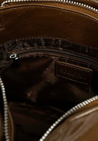 Interior de un bolso de cuero marrón con un forro suave y oscuro, un bolsillo con cremallera y una pequeña etiqueta de cuero con texto en relieve.
