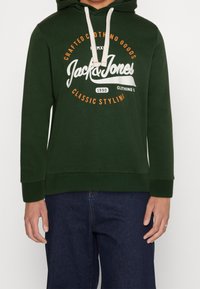 Felpa verde con grafiche frontali, caratterizzata da scritte bianche e arancioni. Coulisse con cappuccio, polsini a costine e vestibilità ampia.