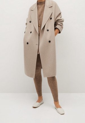 Person trägt einen beigen zweireihigen Wollmantel über braunem Strickpullover, Leggings und weißen flachen Schuhen, steht mit den Händen in den Taschen.