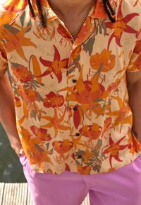 Camicia a maniche corte con bottoni di colore crema, decorata con un vivace motivo floreale arancione e rosso. Presenta bottoni scuri e una vestibilità rilassata.