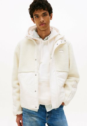 White fleece jacket with quilted lower section. Stylish, layered hoodie underneath with light gray and white tones. Denim jeans complete the look.