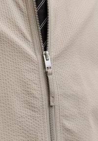 Κοντινή λήψη ασημένιου φερμουάρ μερικώς ανοιχτό σε υφασμάτινη μπεζ υφή, αποκαλύπτοντας μαύρο και λευκό ριγέ ύφασμα από κάτω.