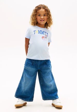 Νεαρό παιδί με σγουρά μαλλιά που φοράει ανοιχτό μπλε μπλουζάκι Tommy, φαρδιά τζιν παντελόνια και λευκά αθλητικά παπούτσια, στέκεται με τα χέρια πίσω από την πλάτη.