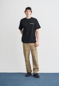 Czarny t-shirt z krótkim rękawem z białym logo „Carhartt”, w połączeniu z beżowymi spodniami bojówkami z bocznymi kieszeniami oraz czarnymi trampkami. Gładkie tło.