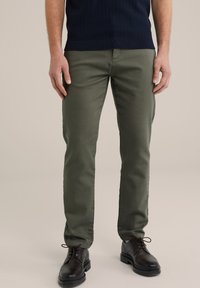 Chinos de algodón verde oliva con diseño de pierna recta, que cuentan con bolsillos y se combinan con zapatos negros con cordones, sobre un fondo neutro.