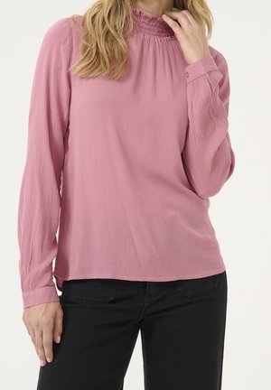 Femme portant un chemisier rose texturé à manches longues avec un col froncé et un pantalon noir, main touchant son cou.