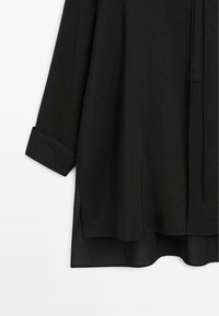 Schwarzes Langarmhemd mit knöpfbaren Manschetten, Seiten-Schlitzen und einem etwas längeren Saum hinten, dargestellt vor weißem Hintergrund.