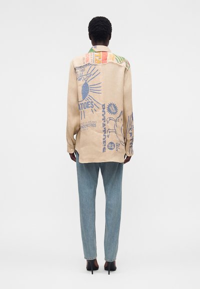 Άτομο που φοράει μπεζ υπερμεγέθη πουκάμισο με μπλε γραφικές εκτυπώσεις και ανοιχτό μπλε τζιν, στέκεται με την πλάτη προς τα εμάς σε απλό φόντο.