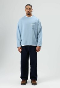 Lichtblauwe oversized sweatshirt met een borstzak, met witte geborduurde tekst, gecombineerd met donkerblauwe spijkerbroek en bruine veterlaarzen.
