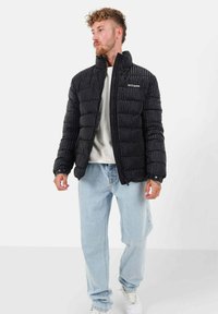 Zwarte pufferjack met een textuur en doorgestikte ontwerp, hoge kraag, voorkant met rits en logo-detailing. Draagt over een lichte sweatshirt en jeans.