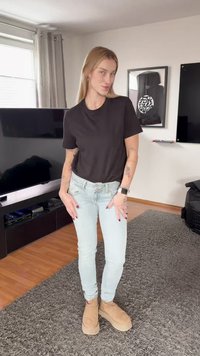 Νέος με μακριά ξανθά μαλλιά, φορώντας μαύρο μπλουζάκι, ανοιχτό μπλε τζιν και μπεζ παπούτσια, στέκεται σε ένα μοντέρνο σαλόνι.