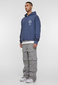Blauwe oversized hoodie met een voorkraag en kruisgrafiek, gecombineerd met lichtgrijze cargobroek met meerdere zakken en witte sneakers.