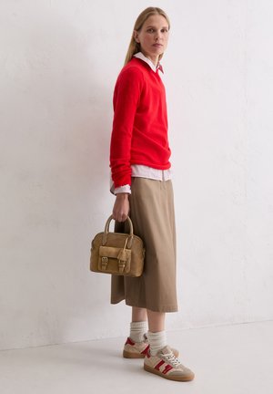 Woman in red sweater and beige skirt holding tan handbag, wearing beige and red sneakers with white socks, standing against a white wall.