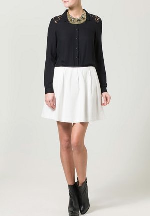 Femme portant un chemisier noir à manches longues, une jupe plissée blanche, des bottines noires et un grand collier doré imposant.