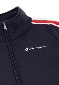 Chaqueta con cremallera color navy, fabricada en una mezcla de algodón, con un cuello alto y detalles a rayas rojas y blancas en los hombros. Logo impreso en blanco.