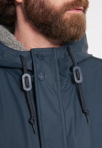 Námořnická voděodolná bunda s šedým límcem z kožešiny, která má černé stahovací šňůrky, patentky a hladký, lesklý povrch.