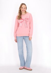 Felpa di cotone rosa con una grande grafica a fiocco bianco e stampa leopardata, abbinata a jeans dritti azzurri e scarpe rosa.