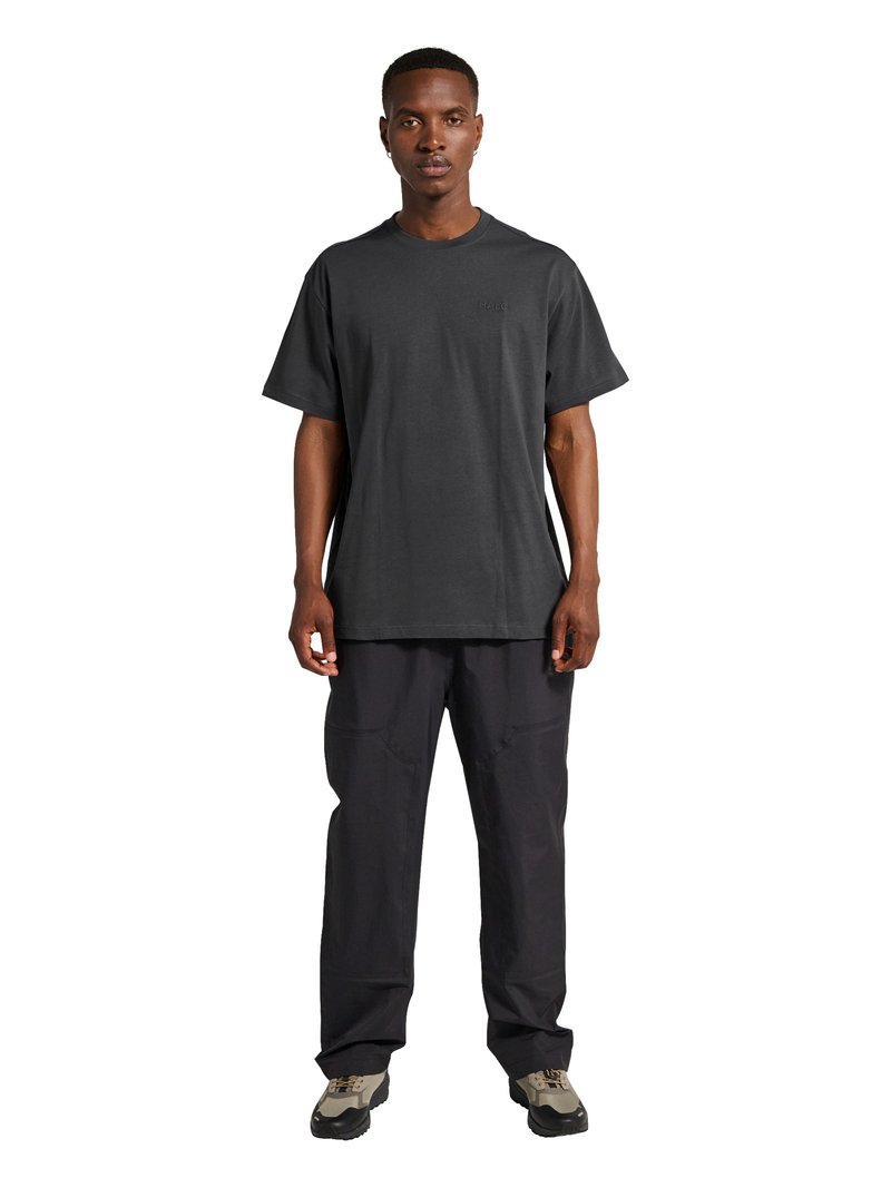 Mężczyzna stojący przodem, ubrany w ciemny, luźny T-shirt, luźne czarne spodnie oraz beżowo-czarne trampki na białym tle.