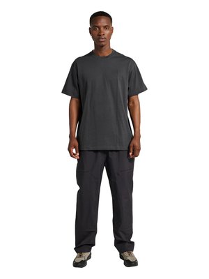 Mężczyzna stojący przodem, ubrany w ciemny, luźny T-shirt, luźne czarne spodnie oraz beżowo-czarne trampki na białym tle.