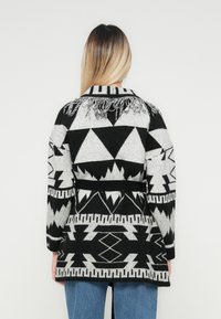 Person med langt blondt hår iført en svart og grå geometrisk mønstrete cardigan med frynser på over ryggen, kombinert med blå jeans.