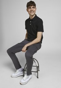 Zwarte polo, grijze broek en witte sneakers. Zittend op een metalen kruk, toont een casual outfit met een eenvoudige stijl en een gladde textuur.