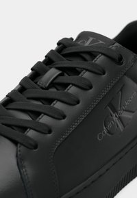 Közelkép egy fekete Calvin Klein sportcipőről, amely részletes varrást, fűzőt fűzőzárral és dombornyomott CK logót mutat az oldalsó paneleten.