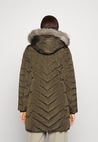 Frau in einer olivgrünen, gesteppten Winterjacke mit einer Fellkapuze, die mit dem Rücken zu einer schlichten Hintergrund steht.