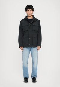 Giacca nera impermeabile con molte grandi tasche, chiusure a scatto e colletto alto. Indossata sopra una maglietta nera, abbinata a jeans chiari e scarpe.