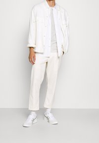 Witte lichte jas over een grijs t-shirt, gecombineerd met crèmekleurige pantalons en witte sneakers met groene accenten. Simpel en casual fit.