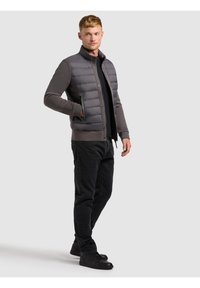 Fiatal férfi rövid hajjal, szürke bélelt dzsekit visel, sötét inggel, fekete farmerral és fekete sportcipővel, egyik kezével a zsebében áll.