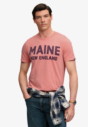 Mężczyzna w różowym t-shircie z nadrukiem "MAINE NEW ENGLAND", niebieskich dżinsach, kratowanej koszuli zawiązanej wokół talii oraz z zegarkiem na rękę.