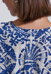 Väljä beige pusero, jossa monimutkaiset siniset kukkakuviot. Napitus takana, pyöreä pääntie ja teksturoitu kangas.