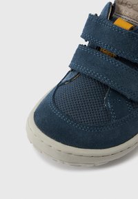 Μπλε ναυτικό αθλητικό παπούτσι με επάνω μέρος από σουέντ και δίχτυ, διαθέτει δύο λουριά με βέλκρο, μπεζ λαστιχένια σόλα και αντίθετες κίτρινες λεπτομέρειες.