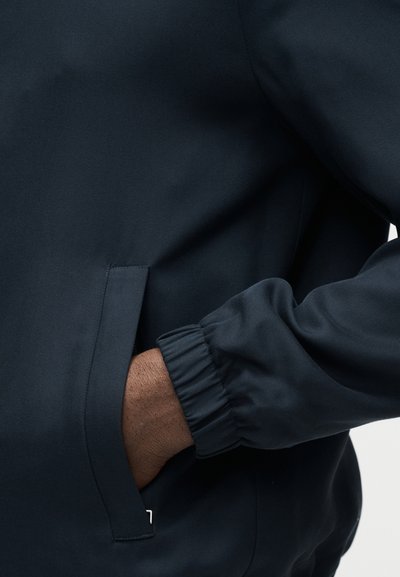 Veste bleu foncé avec poignet élastique sur la manche, main droite partiellement dans la poche avant, tissu lisse et sans plis.