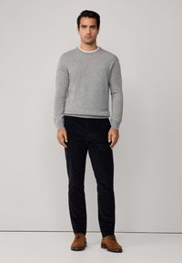 Szary sweter dziany z okrągłym dekoltem, w połączeniu z czarnymi spodniami z kordurowej tkaniny i brązowymi zamszowymi butami. Model stoi na neutralnym tle.