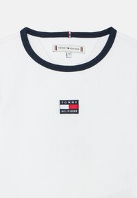 Tricou alb cu dungi mici, cu guler rotund bleumarin. Are un mic logo brodat "TOMMY HILFIGER" în culorile roșu, alb și albastru pe piept.