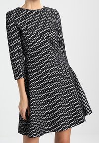 Robe noire à manches trois-quarts, ornée d'un motif géométrique blanc, avec un col rond et une silhouette évasée. Texture de tissu lisse.